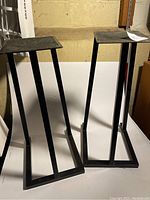 Two black metal speaker stands on white surface against wall background. Both stands have rectangular bases and top platforms, a minimalist design with two vertical supports at the back and a single center vertical support.