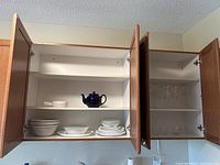 Kitchen cabinets open showing Corelle platter, plates, bowls, blue teapot, and glassware on shelves.
