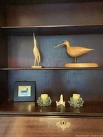 Photo of two wood carved bird sculptures on shelf with framed bird print and two crystal candle holders below.