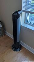 Full view of black and gray Seville Classics tower fan standing upright near a window.