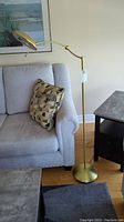 Photo showing full lamp standing next to couch and table, displays lamp structure, base and arm curvature.