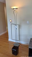 Full view of tall metal framed floor lamp with two lights and dimmer knobs, standing next to a wall on hardwood flooring.