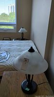 Photo showing one lamp from slightly above, highlighting the frosted glass shade and the metal base beside a bed.