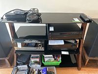Front view of media cabinet showing black glass shelves holding multiple audio devices, two large floor speakers, remote controls on top, and crates of CDs and DVDs below.