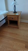 Side view of the wooden nightstand showing the drawer and cubby beneath, placed next to a bed with a lamp on top.