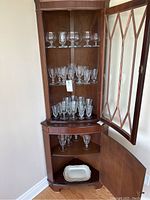 Full view of wooden corner cabinet displaying three shelves of various crystal glassware and an open bottom cabinet with a white ceramic platter