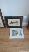 Two framed black and white prints on floor, one leaning against wall (rustic bridge), one laying flat (water scene with trees and animals).
