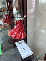 Front view of the glossy porcelain figurine standing on a glass shelf, showing the full flowing red dress and green bonnet.