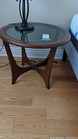Full view of round side table showing dark wood frame, geometric leg design, and glass top insert.