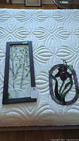 Photo of both sun catchers on a white quilted surface, showing the rectangular glass panel with resin tree design on left and the oval stained glass flower on right.