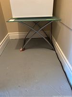 Photo showing frosted glass top table with metal base, viewed in a corner setting, showing whole table and floor space.