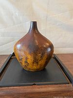 Front view of a large brown pottery vase with a bulbous body and narrow neck on a black tray, showing surface texture and coloration.
