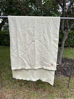 Photo of the off-white wool blanket hanging on a rod outside, showing satin ends and overall condition.