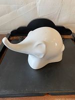 Side view of glossy white ceramic elephant statue with elongated trunk extending left, placed on black tray with wooden frame.