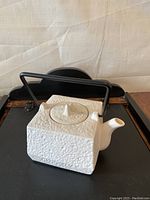 Front side view of a square white ceramic teapot showing textured surface and triangular lid knob with black metal handle above.