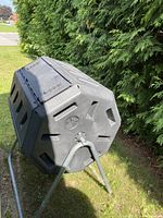 Overall side view of black plastic compost bin on grey metal rotating stand on grass near hedge
