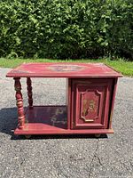 Side view of the pink side table showing turned legs, lower shelf, and cabinet door with gold floral design.
