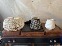 Three lamp shades on a wooden furniture piece, arranged left to right: small white woven shade, black and white geometric shade, large white pleated shade.