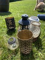 Seven outdoor candle holders of various styles displayed on natural grass with a dog in the background.