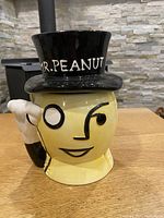 Front and side view of vintage Mr. Peanut ceramic jar with black top hat, yellow faded color, and intact rubber seal.