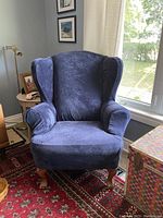 Chair with blue velvet cover showing wingback design, rolled arms, wooden legs, sitting on a red patterned rug near window.