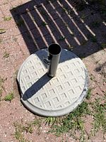 Concrete umbrella base with metal pole and noticeable breakage on the edge, photographed on brick ground with some grass.