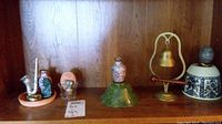Shelf display showing all items in the lot including brass bell with wooden ringer, green stone egg, stone bowls, floral vase, and ceramic wine stopper.