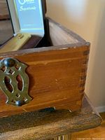 Close-up photo of corner of wooden box showing dovetail joints and brass-colored metal trim, wood surface worn.
