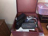 Box with binoculars in black case, camera, glasses case, mirror and other small items inside burgundy storage box