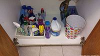 Photo showing various open cleaning supply bottles and sprays organized on a white plastic tray under a sink, plus a polka dot garbage can to the side.