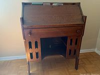 Front view of the wooden desk showing pull-down work surface closed, one drawer below, and decorative cut-out side panels with storage space underneath.