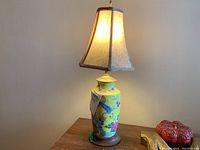 Overall lit lamp showing the floral yellow ceramic base, wooden base, and tan cloth lampshade with brown trim.