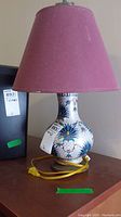 Full view of the lamp showing the red lampshade, ceramic base with colorful floral pattern, and attached yellow power cord.