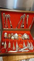 Full view of mixed silver plate flatware pieces inside an old flatware box with orange-red felt lining, showing forks, spoons, knives, and napkin rings.