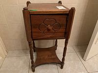 Front angle of wood smokers table showing carved floral design on front door and turned legs, placed on tile floor.