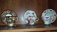 Three tea cups and saucers displayed on wood shelf showcasing floral designs in black with yellow and pink flowers, white with pink and green florals, and white with pink florals with some wear and repair visible on one cup bottom.