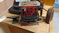 Vintage toaster with red and black ornamental metal housing, power cord wrapped around base, shown on wooden table with other items.