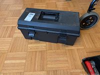Closed black plastic toolbox on wooden parquet floor, showing the outer shell and handle