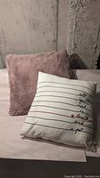 Two cushions on a flat surface against a concrete wall; dusty rose fuzzy cushion and white cushion with black lines and writing.