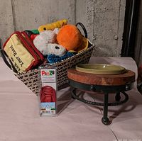 Basket filled with assorted plush dog toys, ceramic Disney brand dog bowl on a wooden and metal stand, and a pack of small size Pawz brand protective dog boots