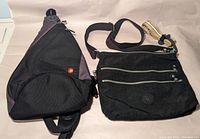 Photo showing one black Swiss Gear sling bag with zippered compartments and one black Kipling crossbody bag with dual zippers and keychain on strap, both bags placed against neutral background.