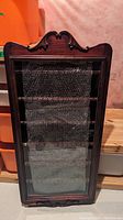 Front view of the wooden display cabinet showing glass door and carved decorative details on top and bottom edges, shelves visible inside.