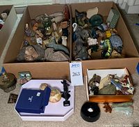 Wide view of cardboard boxes filled with assorted small animal figurines and decorative items, along with small keepsake boxes and one black circular object on floor.