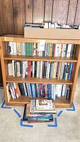 Wooden bookcase with three shelves filled with books and additional books stacked on the floor in front.