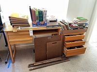Photo showing the small wood desk and the three drawer wood storage unit with multiple binders, books, and art papers stacked on top along with wooden easel parts and artist boards leaning against.
