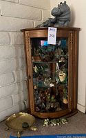Front view of the wooden corner cabinet with glass shelves displaying various animal figurines. Large and small hippopotamus figures on top, solid brass turtle and three green frogs on floor beside it.