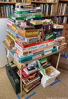 Image showing a large stack of board games of various titles piled on and under a table with bookshelves in the background.