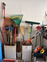 Wide view of two tall containers holding various long handled garden tools like rakes, brooms, shovels, plus electric power tools (weedwhacker, hedge trimmer, chainsaw) on ground around them.