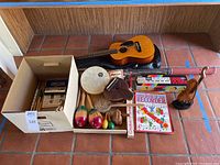 Wide shot showing acoustic guitar with soft case, percussion instruments including maracas, tambourine, gourd piano, recorder, instruction book, and Casio VL-Tone keyboard box