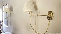 Pair of brass wall lamps mounted on white wall, showing pleated cream fabric shades and yellow cords hanging down